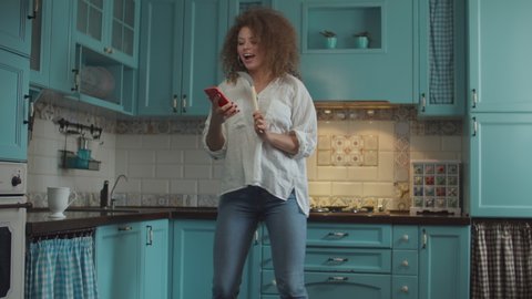 Smiling curly 20s woman in jeans dancing happily with scoop as microphone and mobile phone in hands on home blue kitchen. 