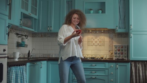 Smiling curly 20s woman in jeans dancing happily with scoop as microphone and mobile phone in hands on home blue kitchen. 