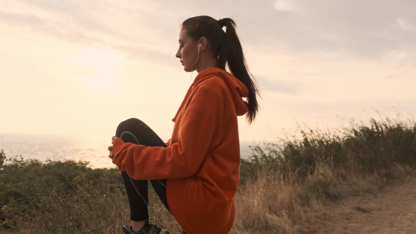 Side view of Concentrated pretty athletic woman in earphones warming up with legs and looking away at seashore Royalty-Free Stock Footage #1048124752