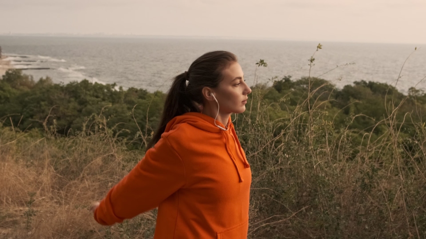 Carefree pretty athletic woman in earphones warming up with arms and looking away at seashore Royalty-Free Stock Footage #1048124857
