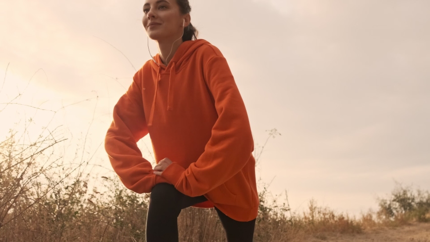 Happy pretty athletic woman in earphones warming up with legs and looking away at seashore Royalty-Free Stock Footage #1048124866