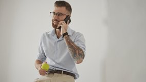 A peeping view of a positive young man in eyeglasses is holding a tennis ball while talking on the phone standing in the white office