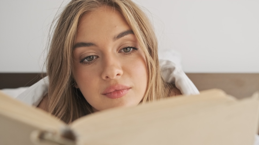 A close-up view of a smiling positive young woman is reading a book while lying in the bed in the morning | Shutterstock HD Video #1048125076