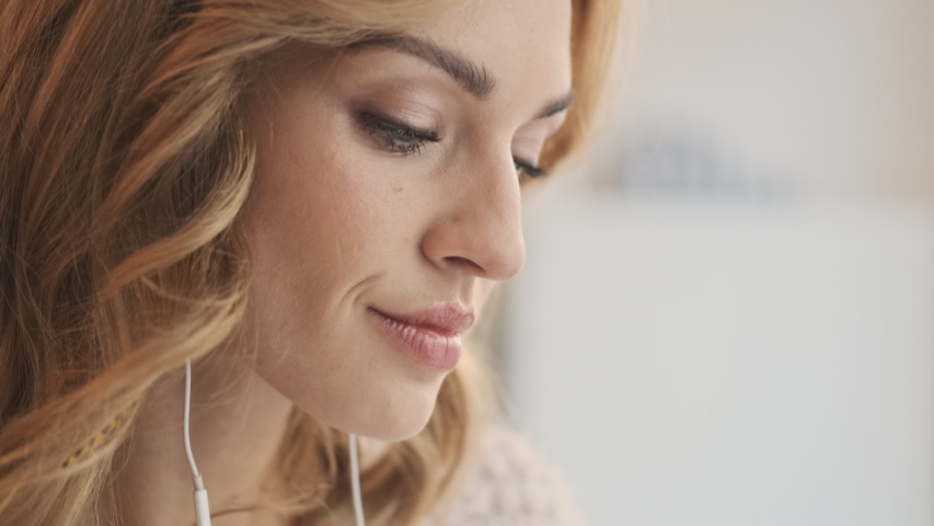 A close-up view of a happy lovely nice blonde woman is watching something on her smartphone and laughs while sitting in the white cozy cafe