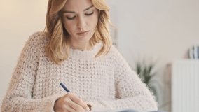 A focused young blonde woman is writing exercise in a cozy place indoors
