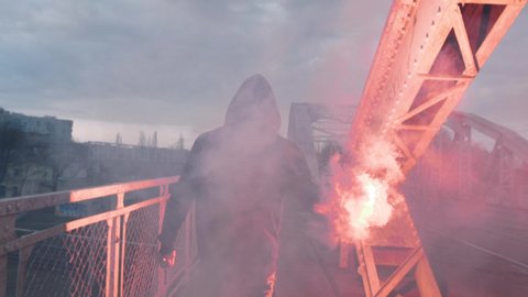 Back view of a young man in hoodie and balaclava with red burning signal flare on the road under an old steel frame bridge, slow motion