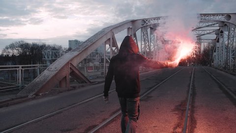 Young man in hoodie and balaclava with red burning signal flare on the road under an old steel frame bridge, slow motion