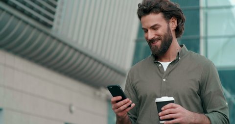 Bearded Handsome Man having a Slow walk in the Town, enjoying his Tasty Coffee. Typing Messages on his Smartphone and drinking hot Coffee. Wearing stylish clothes. Having nice Hairstyle.