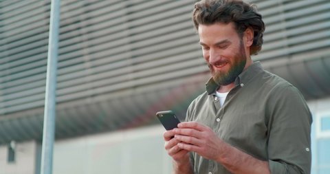 Close up of Attractive Man Using his Smartphone and looking with interest on Screen. Having nice Appearance and stylish Clothes. Handsome man with Well-groomed Hairstyle and Beard.