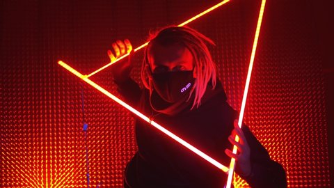 UFA/RUSSIA - 02.03.2020: Young beautiful man model in neon lights. Neon triangle in a club studio. Concept of dubstep or punk style. Model in a red neon studio. Wearing a medical black mask