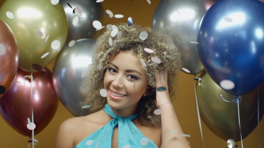 Beautiful hispanic woman in turquoise dress dancing on yellow background with multicolored balloons and flying confetti in slow motion. Pretty mixed race girl celebrates holiday at summer party