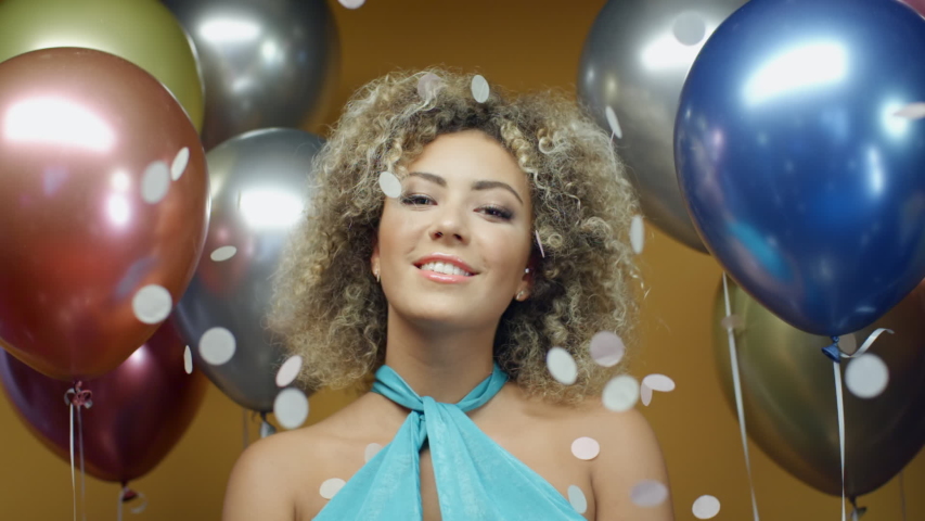 Beautiful hispanic woman in turquoise dress dancing on yellow background with multicolored balloons and flying confetti in slow motion. Pretty mixed race girl celebrating holiday at summer party