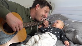 An attractive father playing guitar to his adorable baby boy on a bed. Baby listening and smiling. 4K - Powered by Shutterstock - Get 15% off with code: PIKWIZARD15