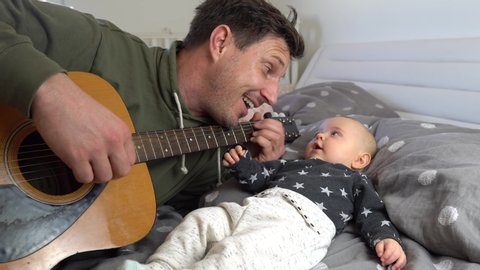 An attractive father playing guitar to his adorable baby boy on a bed. Baby listening and smiling. 4K