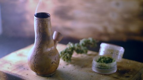 hand puts a smoking bong with marijuana on the table on which there is a grinder and a large bud of medical cannabis.