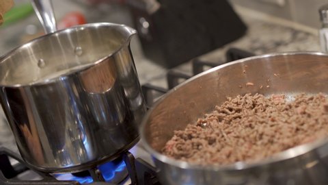 Pouting Pasta in Pot with Ground Beef. a close up view of ground beef cooking in a pan and someone pouring pasta in pot on a stove top