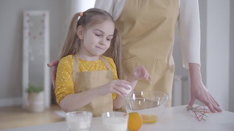 Portrait of little concentrated Caucasian girl breaking egg into bowl. Cheerful child in apron helping grandmother to bake pancakes for Shrovetide. Cooking, lifestyle, Shrove Tuesday.