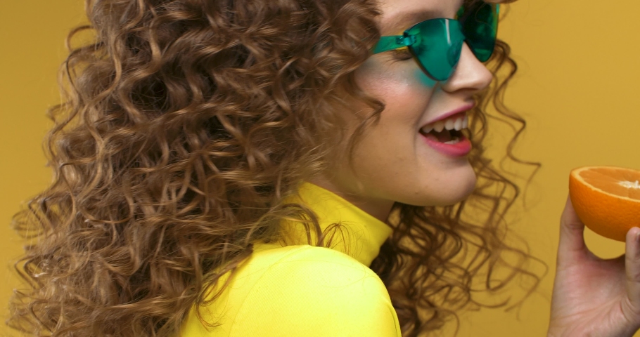 Portrait of Female beautiful Model posing on Camera while Holding part of Sweet Orange. Looking stylish and having trendy Eyeglasses. Neon yellow Top on Creative Woman. Bright Makeup.