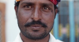 Closeup of a happy cheerful positive young brown man with mustache as he smiles looks straight at camera photo video, adjusts his multicolor vibrant turban, outdoors exterior of his small town house