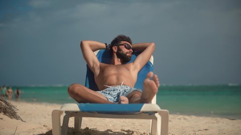 Tan Man Relaxing On Vacation Caribbean Tropical Beach. Tanned Man Lies On Sunbed On Vacation. Summer Sunbathing Resort Sea Or Ocean. Happy Traveler Resting Beach Hotel Luxury. Guy Lying On Sun Lounger