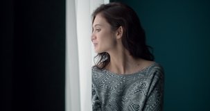 Pretty pensive girl with brown hair standing near window and looking at camera with dramatic mood. Young girl in casual clothing lost in her thoughts at home.