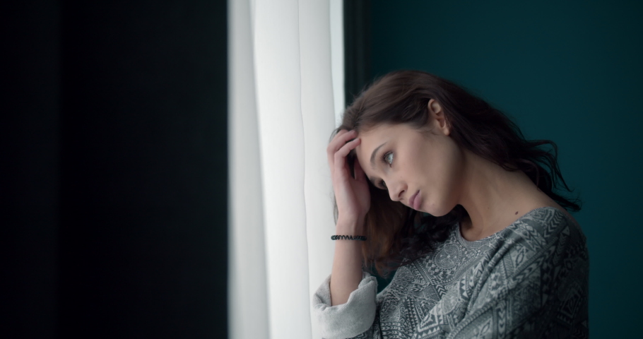 Side view of young thoughtful lady with curly hair dressed casually looking through window at home. Pretty woman feeling loneliness and sadness during leisure time. | Shutterstock HD Video #1048165174