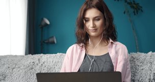 Pretty young lady with dark curly hair in casual clothing sitting comfortably on couch, listening music on headphones and working on laptop. Concept of multitasking and human skills