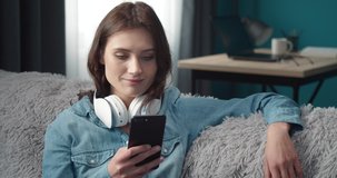 Cheerful young lady in casual clothing sitting on grey sofa with wireless earphones on neck and using smartphone. Smiling woman with curly hair checking her playlist on mobile.