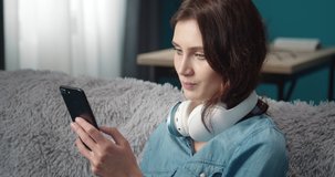Smiling stunning girl with dark curly hair in denim shirt looking at smartphone screen with wireless earphones on neck. Concept of relaxation and enjoyment