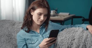 Smiling young lady with brown curly hair sitting comfortably on grey sofa and using smartphone. Happy girl in casual clothing spending free time with mobile at living room.