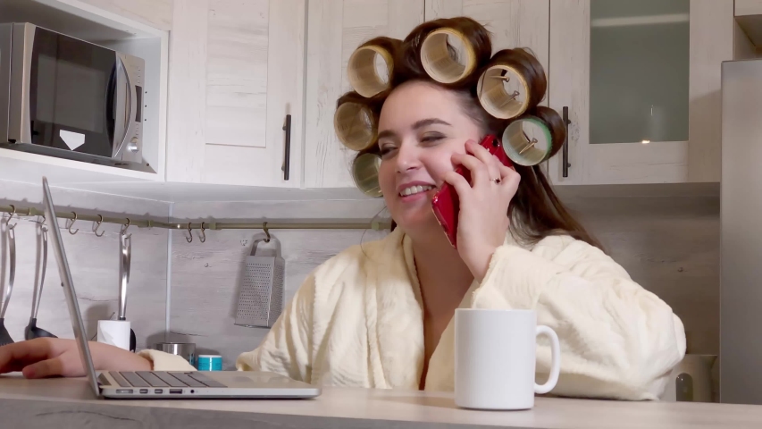 Plus-size girl, dressed in a bathrobe, curlers on her head, talking by phone sitting at kitchen, laptop on desk