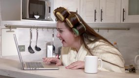 Plus-size girl, dressed in a bathrobe, curlers on her head, talking by video call using laptop at kitchen