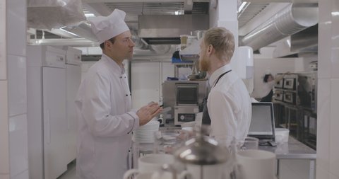 Side view of chef cook in uniform and cap giving instruction about dish serving to young handsome waiter in black apron working together in restaurant kitchen