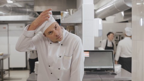 Portrait of exhausted head chef putting off hat and unbuttoning uniform jacket feeling tired after long day at work in restaurant kitchen. Tired chef cook taking break to relax at work in kitchen