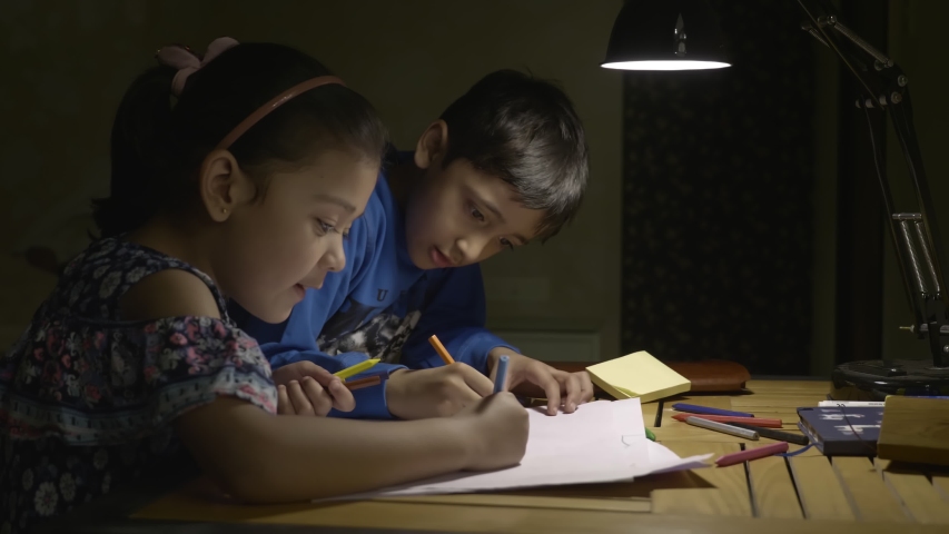 Two young kids drawing on a white paper under a table or desk lamp in the night. A boy and a girl are focused while studying together in the study room.  Royalty-Free Stock Footage #1048229983