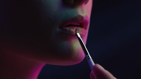 Young Woman Applying Lipstick on Lips. Cosmetics for Make-Up and Gloss of Posing Girl in Multi-Colours of Neon. Close Shot of Modern Makeup on Black Background. Flickering Lighting of Fashion Studio
