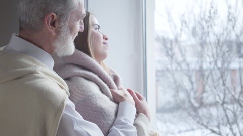 Side view of elegant senior Caucasian man covering young girlfriend with blanket. Portrait of happy couple with age difference looking out the window. Joy, lifestyle, love, care.