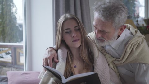 Senior Caucasian man in love talking with young pretty Caucasian woman reading book. Happy loving couple with age difference enjoying time together indoors. Lifestyle, love, romance, tranquility.