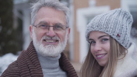 Close-up faces of young woman and senior man looking at each other and at camera and smiling. Happy Caucasian couple with age difference standing on winter day outdoors. Leisure, lifestyle, love.