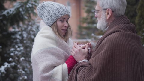 Mature Caucasian man coming to young girl with tea cup standing outdoors and taking her hands. Portrait of adult couple with age difference chatting on winter day. Lifestyle, relationship, leisure.