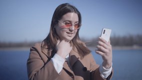 Close-up of joyful young woman in sunglasses talking at selfie camera on smartphone outdoors. Positive smiling Caucasian girl having video call as resting on river bank on sunny autumn day. Lifestyle.