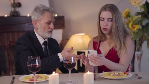 Senior elegant Caucasian man in suit talking to young woman using smartphone. Busy girl in red dress ignoring older partner during romantic dinner. Relationship of couple with age difference.
