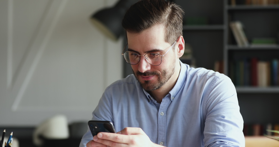 Smiling young man using smartphone indoors. Millennial businessman mobile technology user working in digital applications gadget searching information online, texting messages at home or in office. | Shutterstock HD Video #1048239457