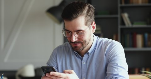 Smiling young man using smartphone indoors. Millennial businessman mobile technology user working in digital applications gadget searching information online, texting messages at home or in office.