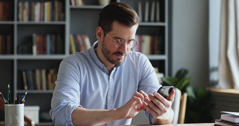Male millennial professional holding modern smartphone texting message in office. Young businessman using helpful mobile apps for business time management organization concept sitting at work desk.