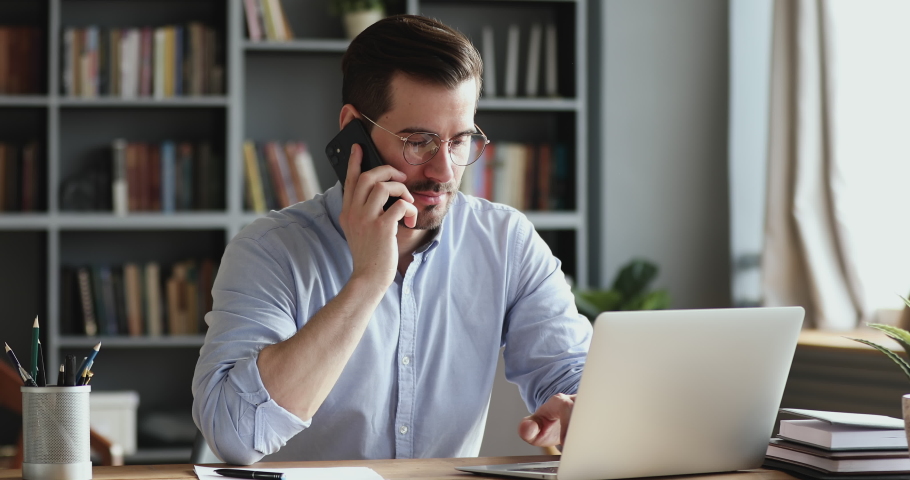 Smiling business man using laptop talking on cell phone sits at desk. Happy confident male professional manager web designer consulting client about online project making business call at workplace. Royalty-Free Stock Footage #1048239469