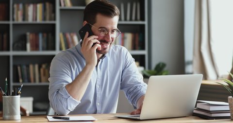Smiling business man using laptop talking on cell phone sits at desk. Happy confident male professional manager web designer consulting client about online project making business call at workplace.