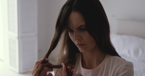 Worried young woman holding brittle fragile hair looking damaged and dry. Unhappy annoyed girl feeling upset about unhealthy split ends having hormone problems or vitamins deficiency concept.