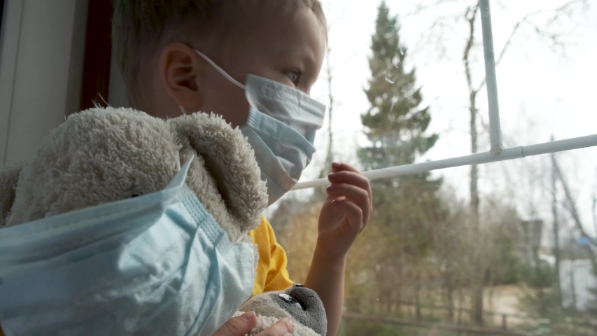 Quarantine, threat of coronavirus. Sad child and his teddy bear both in protective medical masks sits on windowsill and looks out window. Virus protection, pandemic, prevention epidemic. Royalty-Free Stock Footage #1048244092