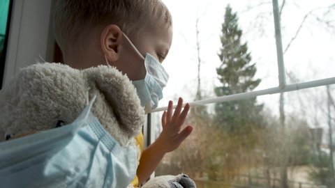 Quarantine, threat of coronavirus. Sad child and his teddy bear both in protective medical masks sits on windowsill and looks out window. Virus protection, pandemic, prevention epidemic.